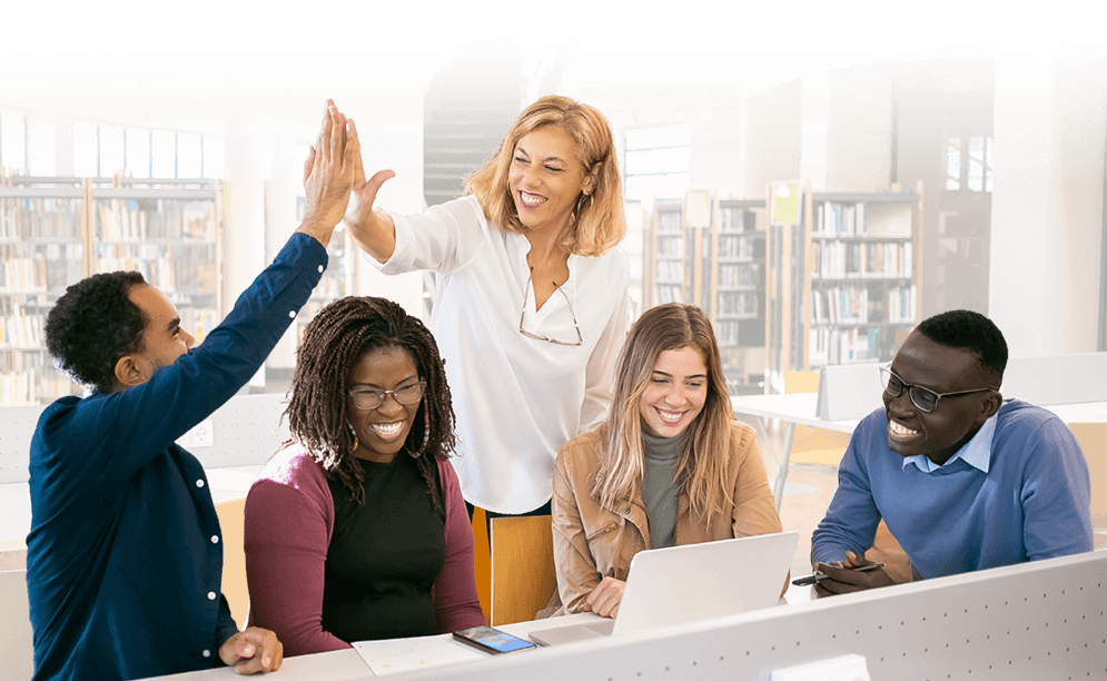 Admin-second-banner-tablet A group of educators sitting around a laptop, smiling, and two are high fiving
